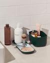 A Small Dark Green Storages Bins with beauty products in it next to a Dark Green Nesting Tray on a white background.