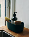 A Small Dark Green Storages Bins with dishwashing supplies in it on a wooden surface.