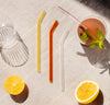 Over head shot of the Poketo Glass Straws in Warm Set on a white background next to a glass of drink and lemons. .