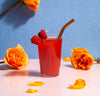 A straw from the Glass Straws in Warm Set in a glass of raspberry juice with orange rosed in the background.