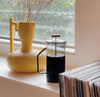 A Gray Glass French Press filled with coffee on a window sill with a yellow vase behind it.
