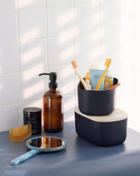 Small Navy Storages Bins with beauty products and one with a  wooden lid on top of each other on a cream background.