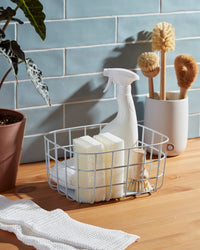 A Open Spaces Light Blue Medium Wire Basket with cleaning supplies on a wooden surface.