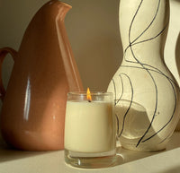 A 8 Oz candle on a cream background with pots behind it.